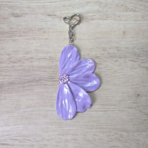 Charm de bolso con forma de media flor, compuesta de 5 pétalos en color lila con detalles iridiscentes El enganche tiene forma de corazón y es en color plateado