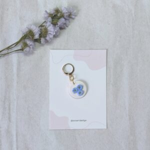 Marcador para agujas o ganchillo, con forma circular en color blanco que incluye tres pequeñas florecillas en azul con el centro en bolitas doradas